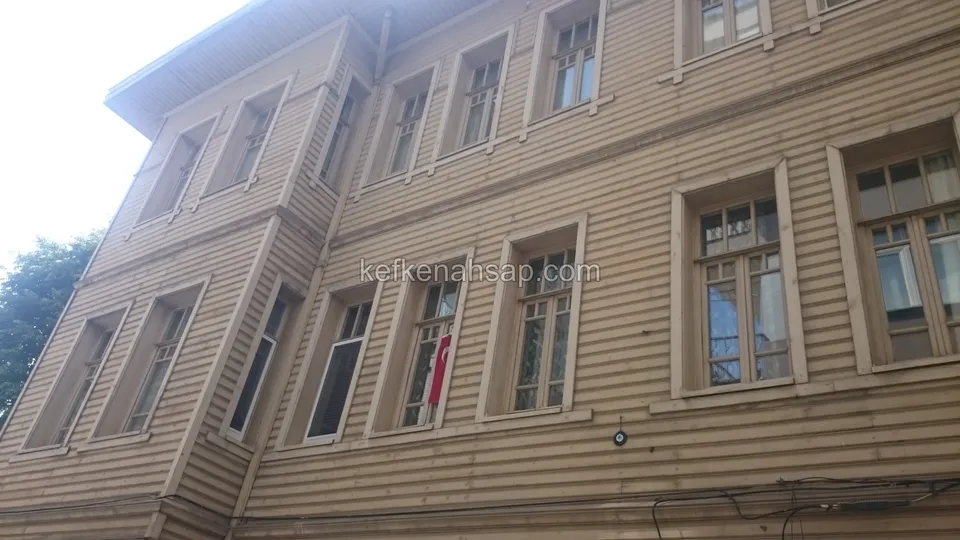 Bej boyalı yatay ahşap kaplamalı, çıkmalı köşesi ve sıra sıra pencereleri olan eski tip bir bina cephesi