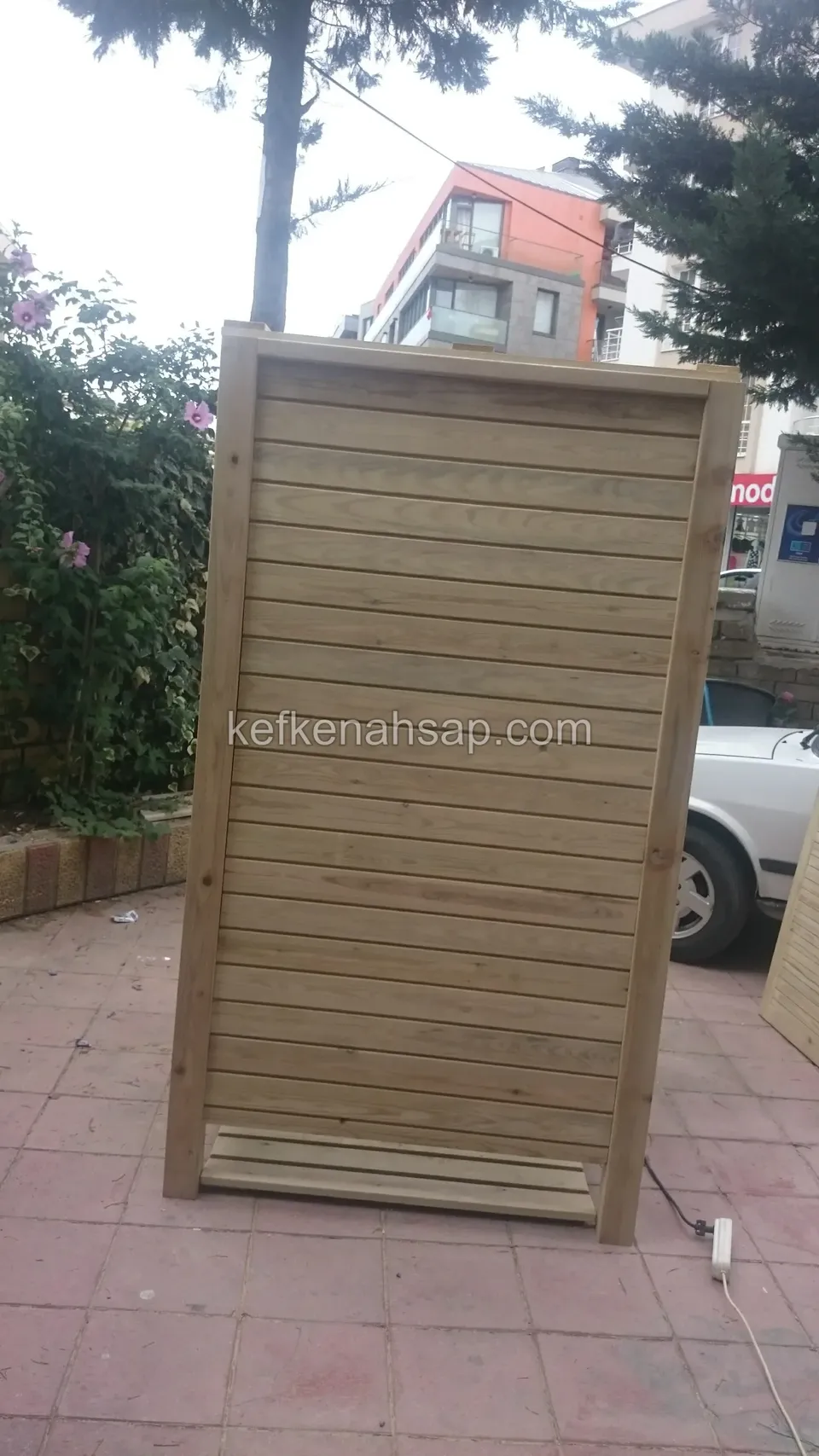 Teras karosu üzerinde duran, yatay tahtalı ve kapalı görünen yüksek ahşap kasa, arkada araç ve ağaçlar