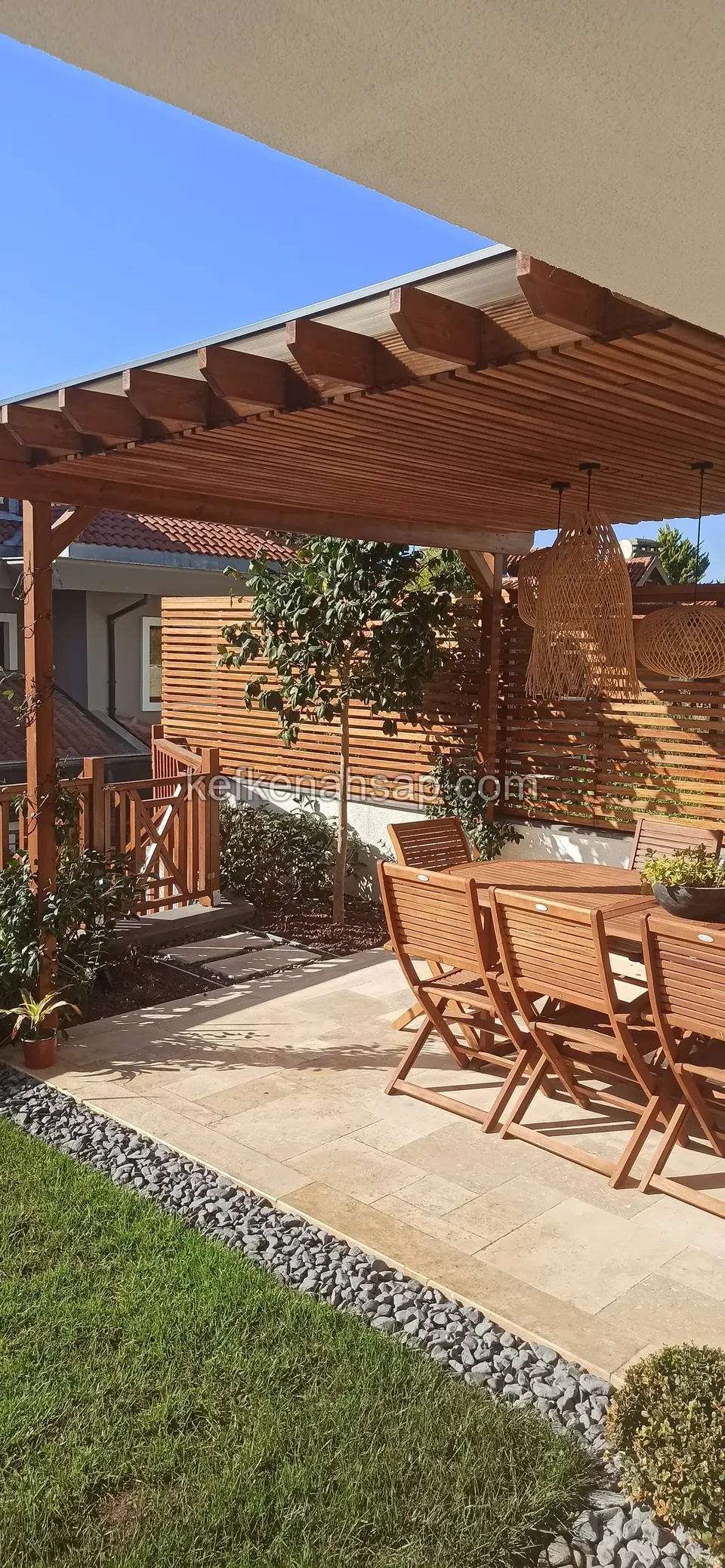 Taş zeminli bahçe terasında, altında ahşap yemek takımı ve hasır avizeler olan ahşap pergola