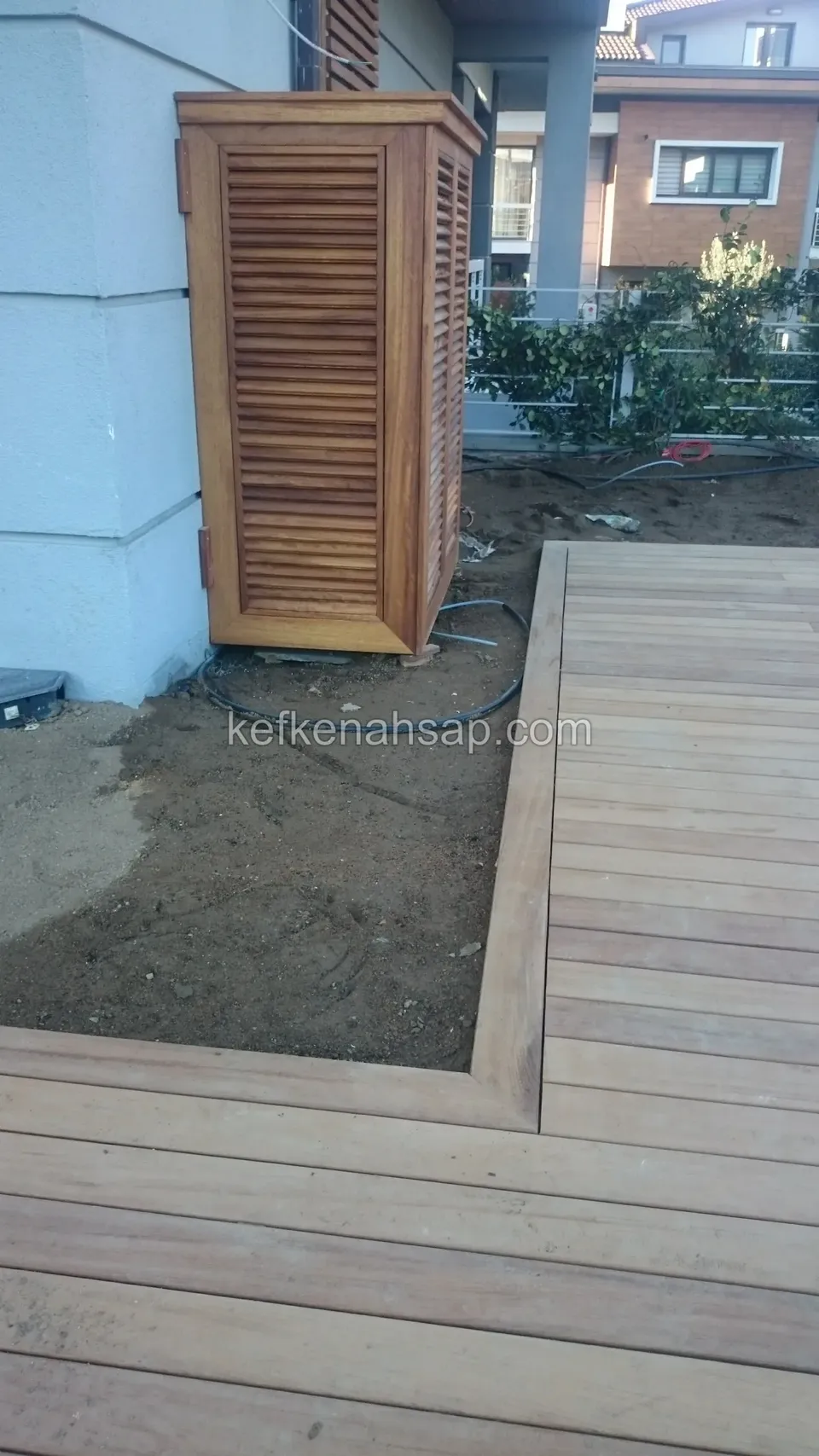 Edge of a newly laid timber deck with a louvred timber cabinet beside it and neighbouring houses behind