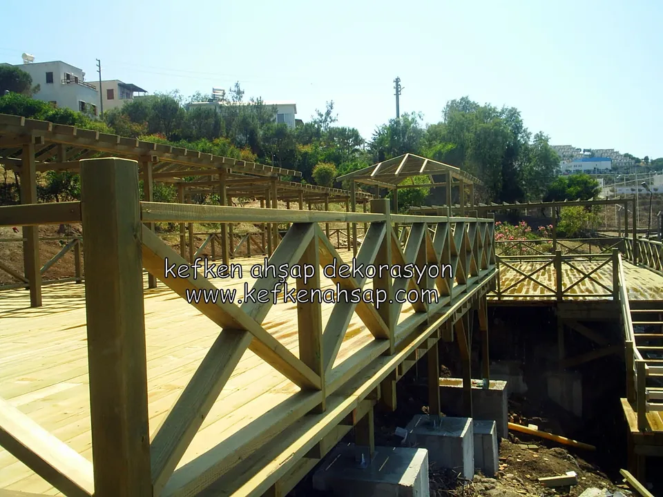 Taze döşenmiş açık renk deck üzerinde kalın dikmeli, çapraz çıtalı korkuluk; arkada pergola ve kıyıdaki yapı