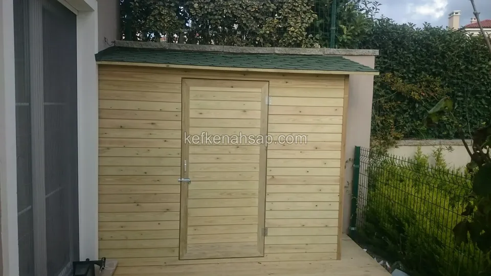 Yatay tahtalı, tek kapılı küçük ahşap depo; üstü yeşil shingle ile kaplı, yanında çit ve çalılık var