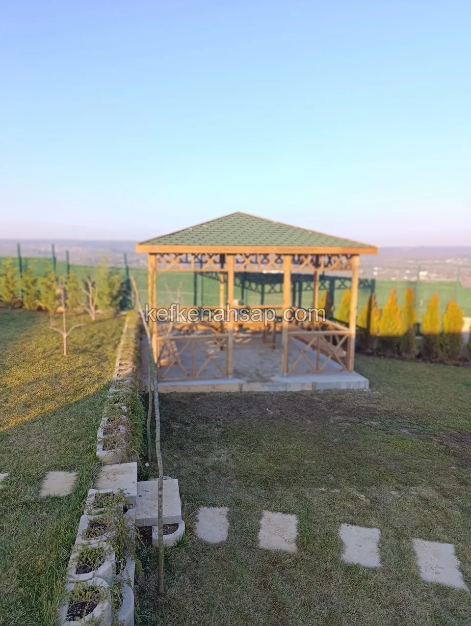 Yeşil çatılı ahşap kamelya, çevrede çimen, beton çiçeklik blokları ve taş adımlar, uzakta ovada çit