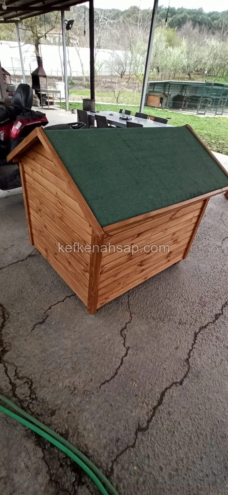 Yeşil kaplamalı çift eğimli çatısı ve bal rengi yatay ahşap kaplaması olan köpek kulübesi, kapalı bir bahçe alanında duruyor