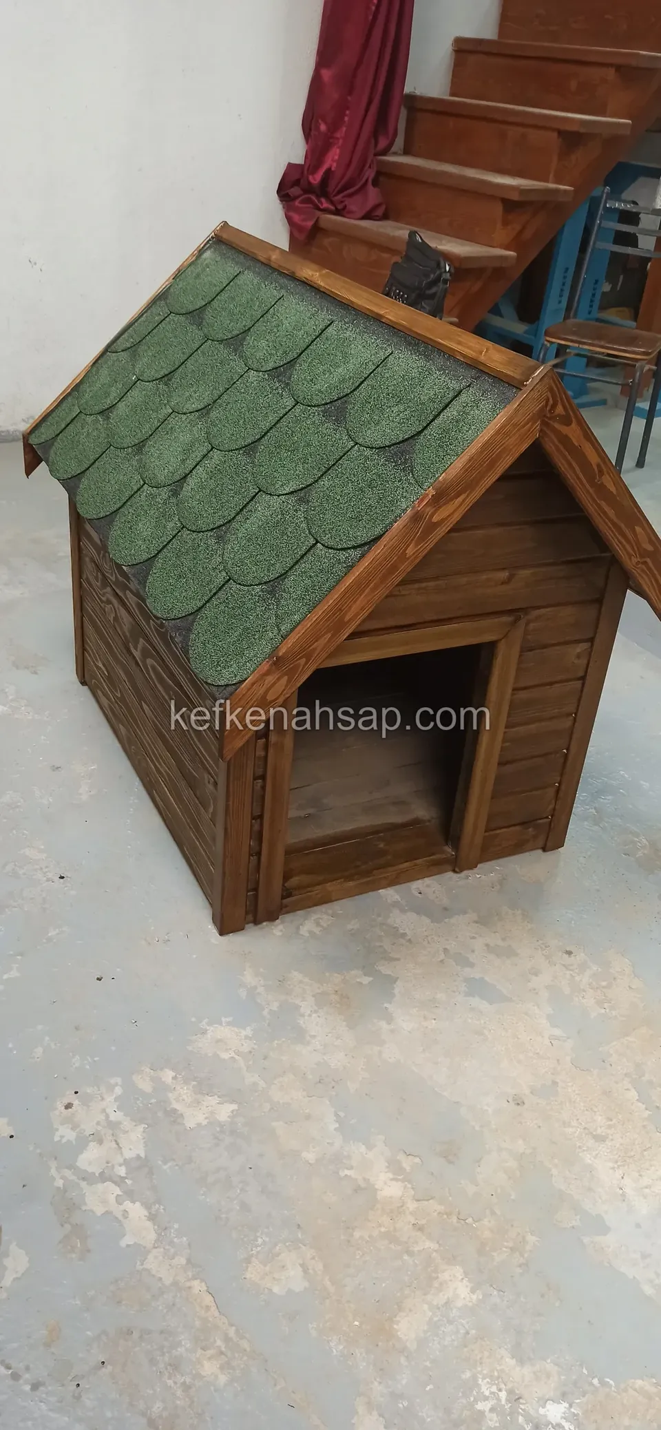 Atölye zemininde çapraz açıdan görünen yeşil çatılı, vernikli ahşap köpek kulübesi
