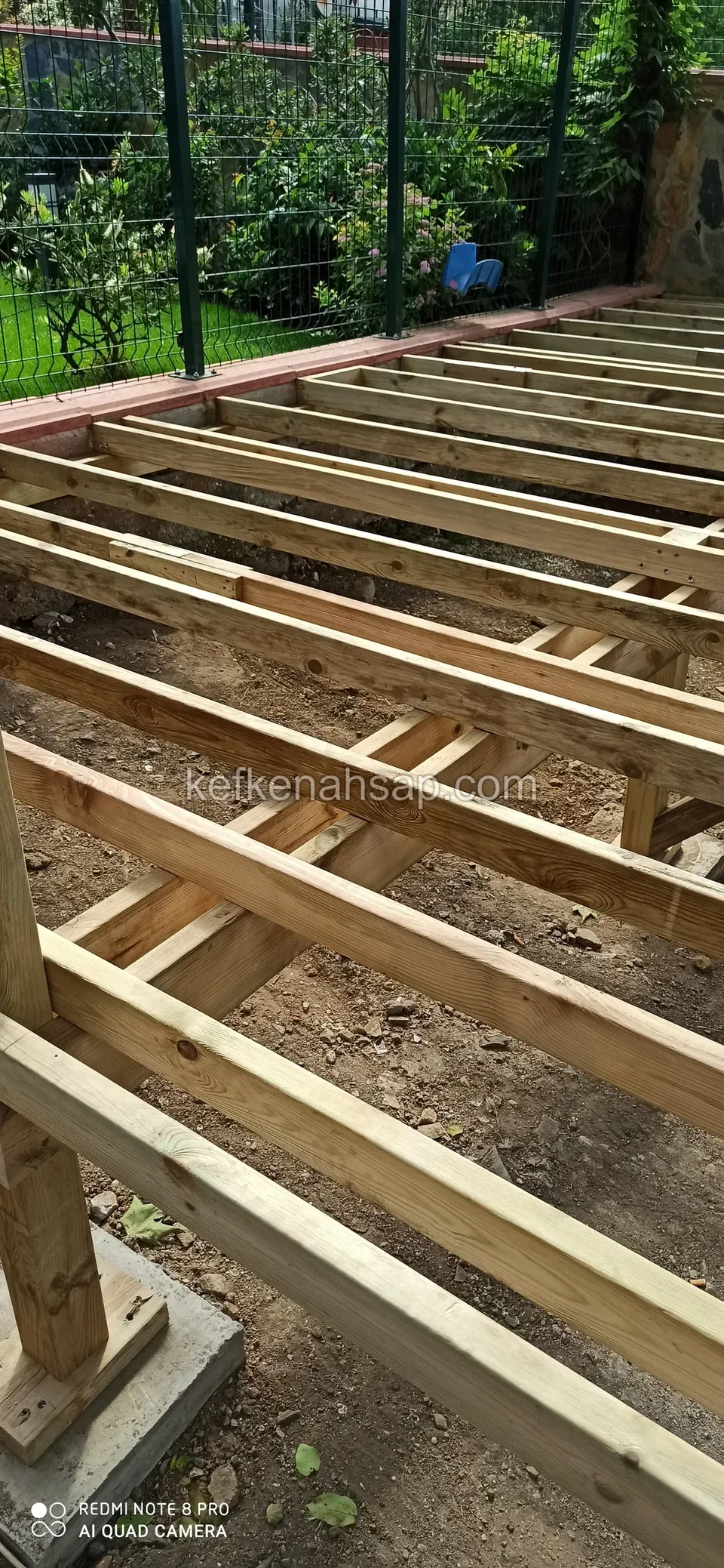 A post on a concrete footing and raised deck joists, with a green fence and garden behind