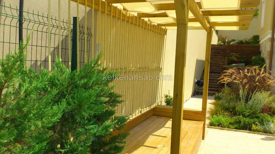 Yeşil telli çit boyunca bahçede pergola direği, dikey çıtalı panel, basamaklı yeşillik ve süs otları