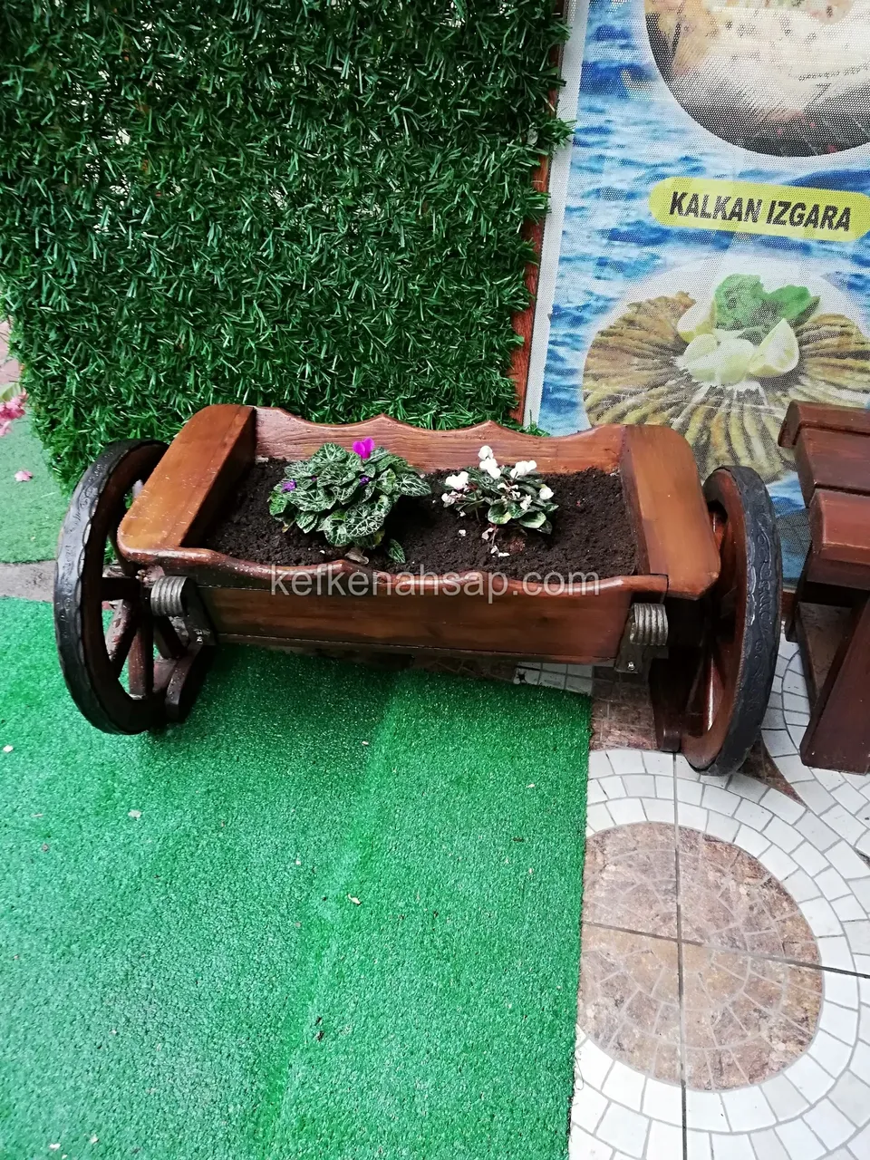 Small timber wheelbarrow with pink and white flowers on green matting, with a colourful poster behind