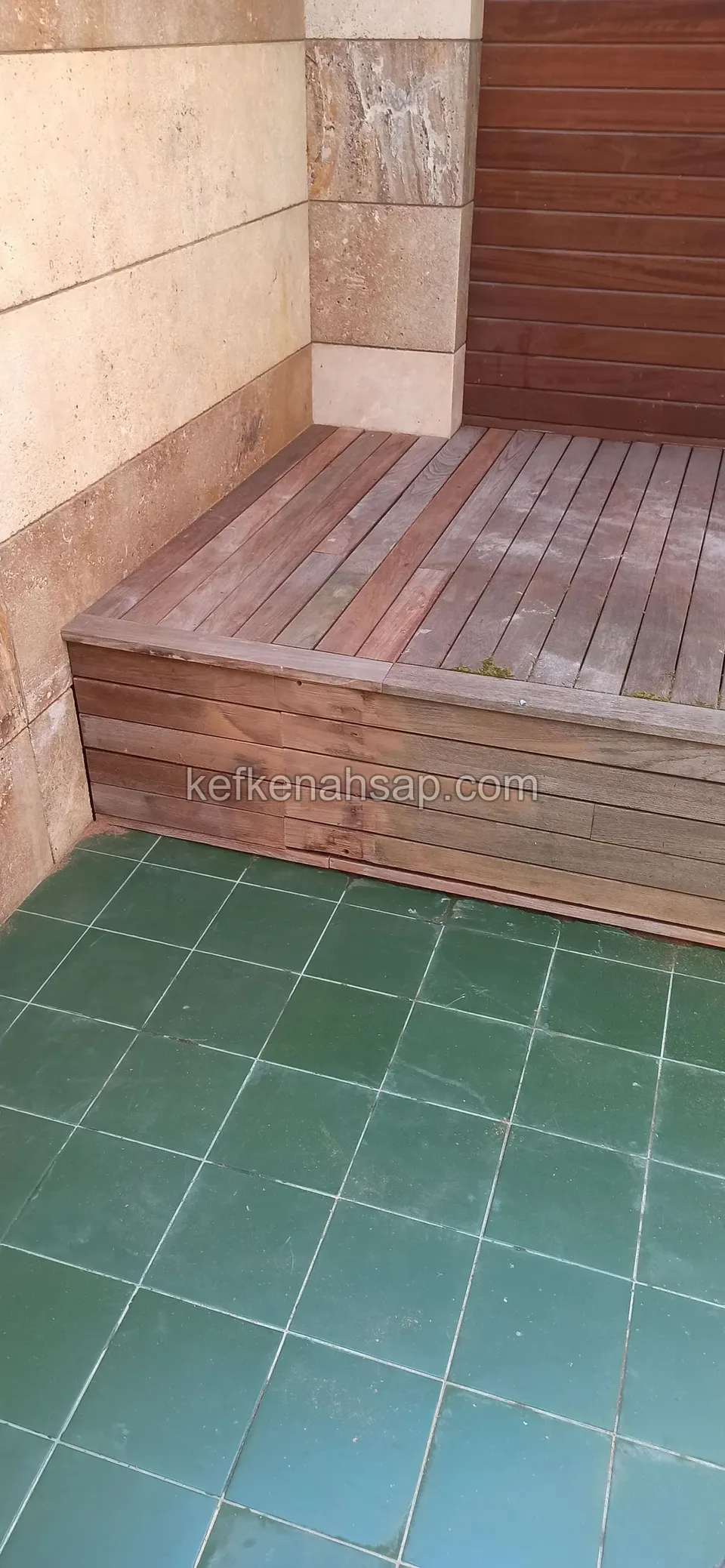 Worn raised timber platform in a corner on a green-tiled floor, with timber wall cladding behind