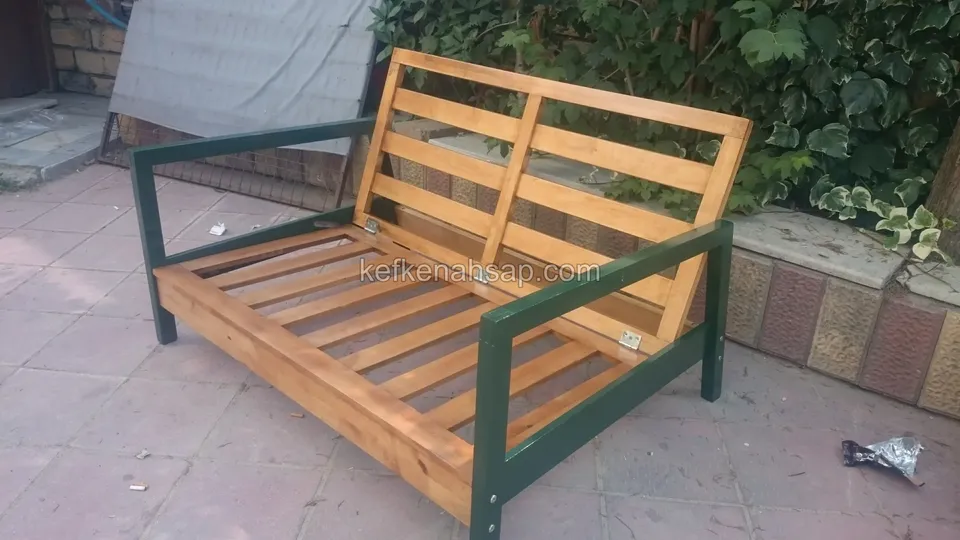 Das Gestell eines zweisitzigen Sofas aus Holzlatten ohne Kissen, mit grün gestrichenen Armlehnen, auf Steinboden