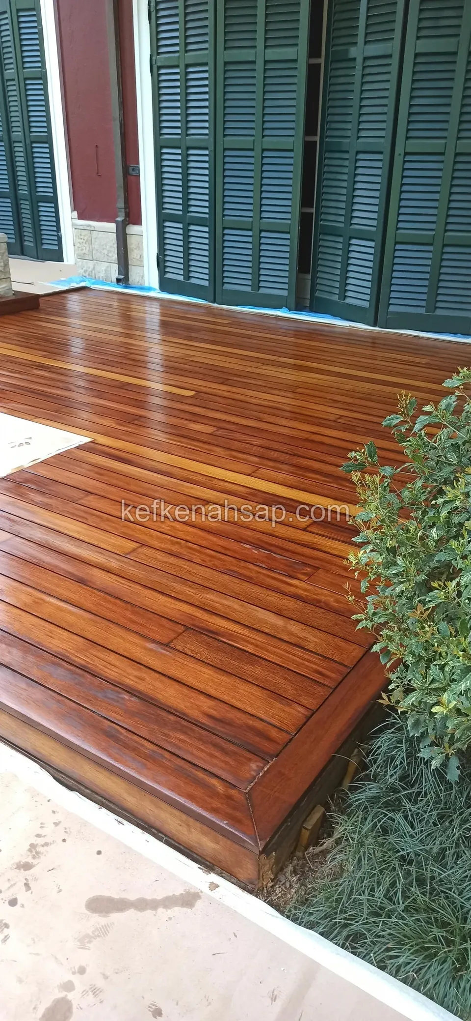 Glossy dark red-brown deck laid in front of green-shuttered doors, edges protected with blue tape