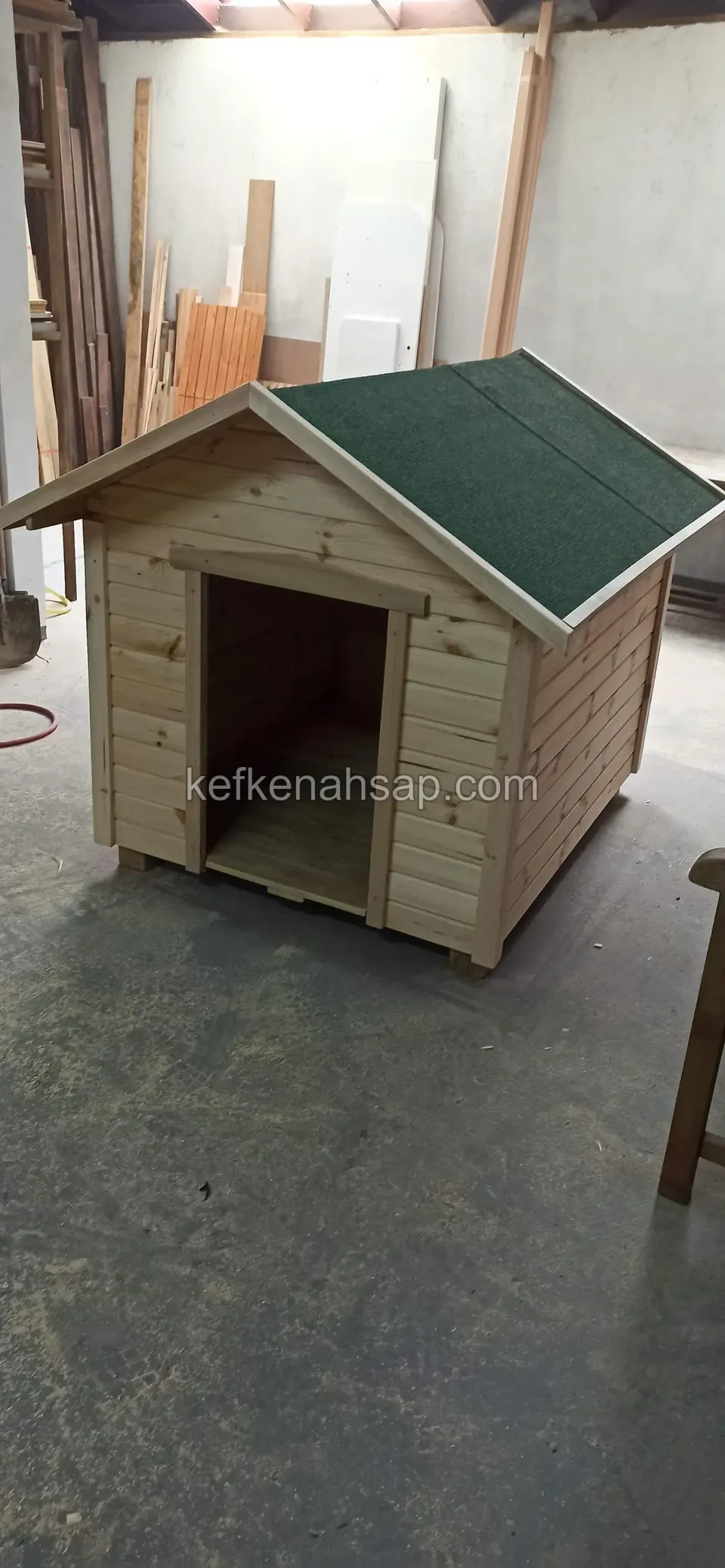 Yeşil shingle çatılı, boyasız açık renk ahşap köpek kulübesi; arkada atölyenin kereste istifi