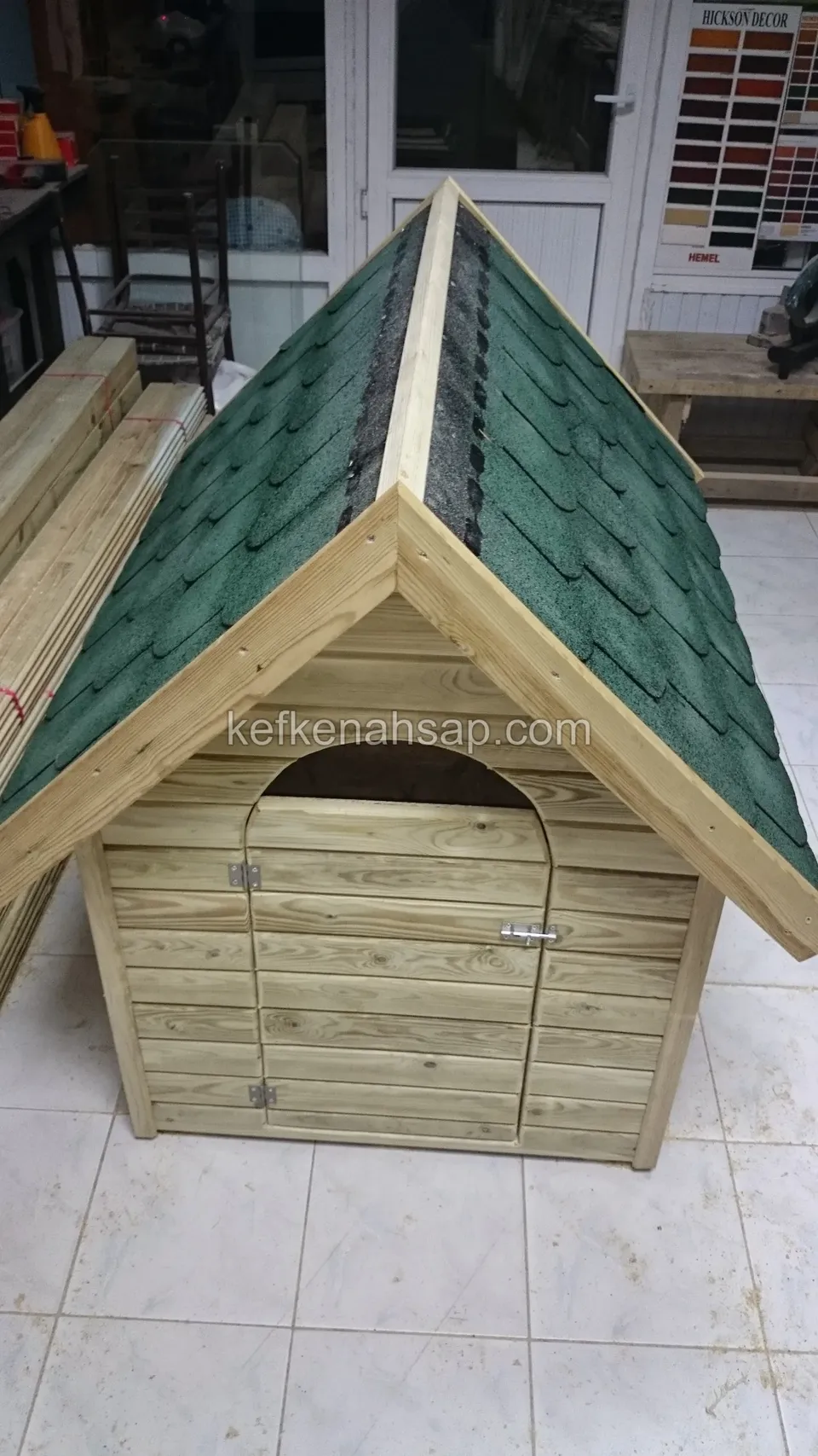 Yeşil shingle çatılı, kemerli kapısı kapalı açık renk ahşap köpek kulübesi; seramik zeminde