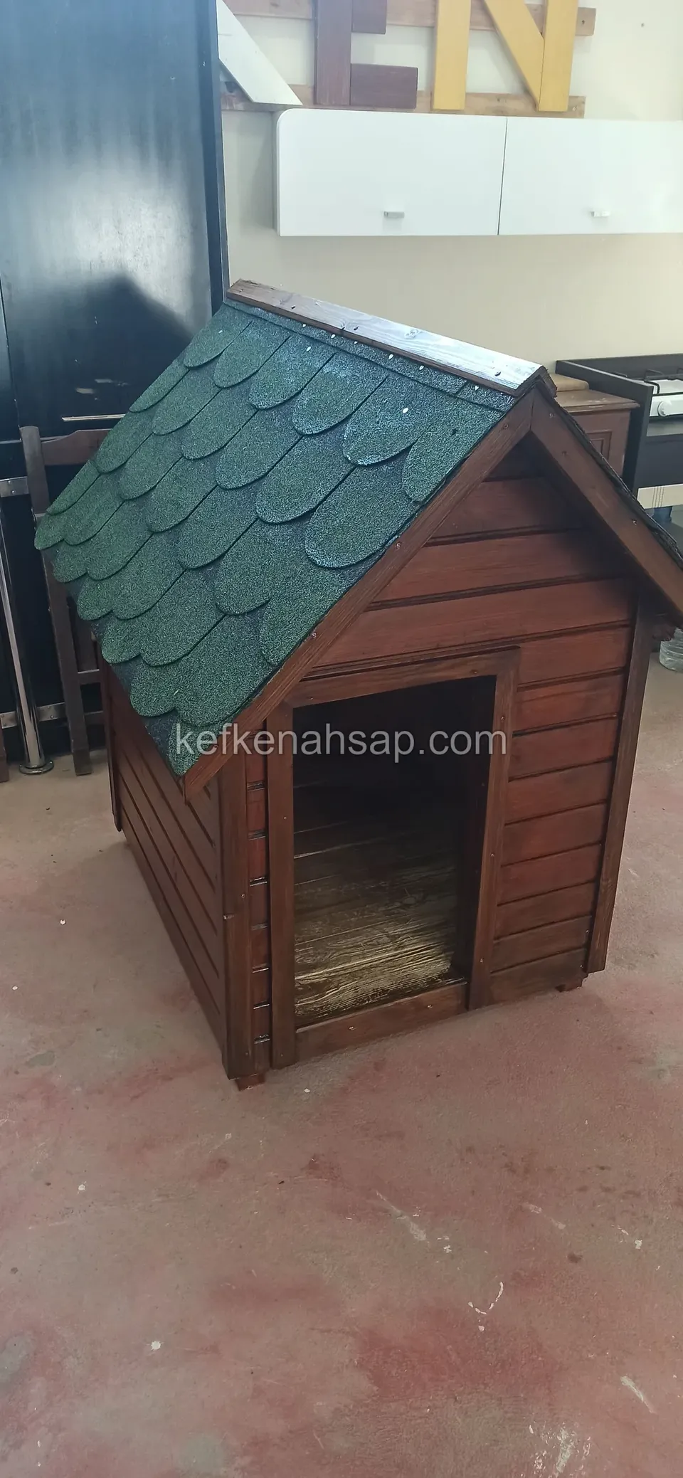 Atölye zemininde duran, yeşil shingle çatılı ve koyu bal rengi vernikli ahşap köpek kulübesi