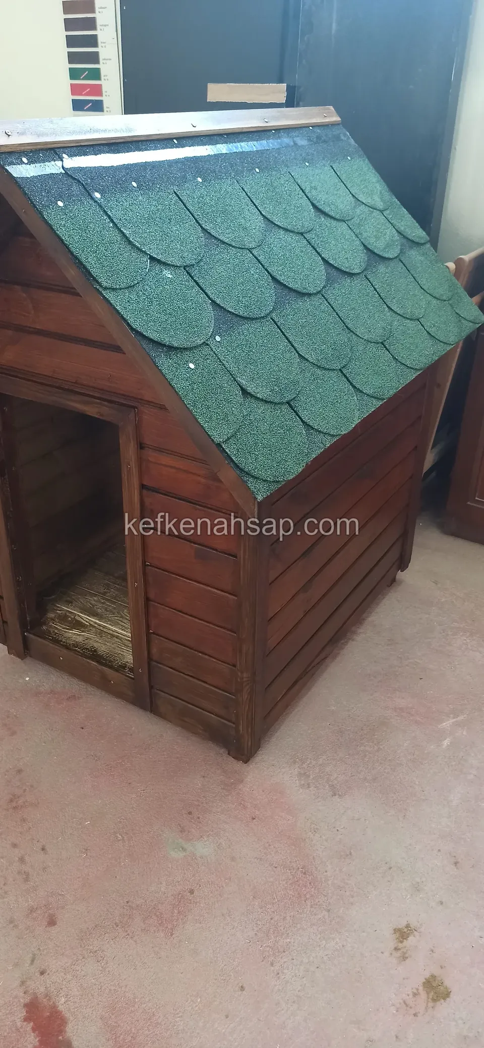 Yatay tahta kaplamalı, yeşil shingle çatılı vernikli köpek kulübesi; yan yüzünde giriş açıklığı görünüyor
