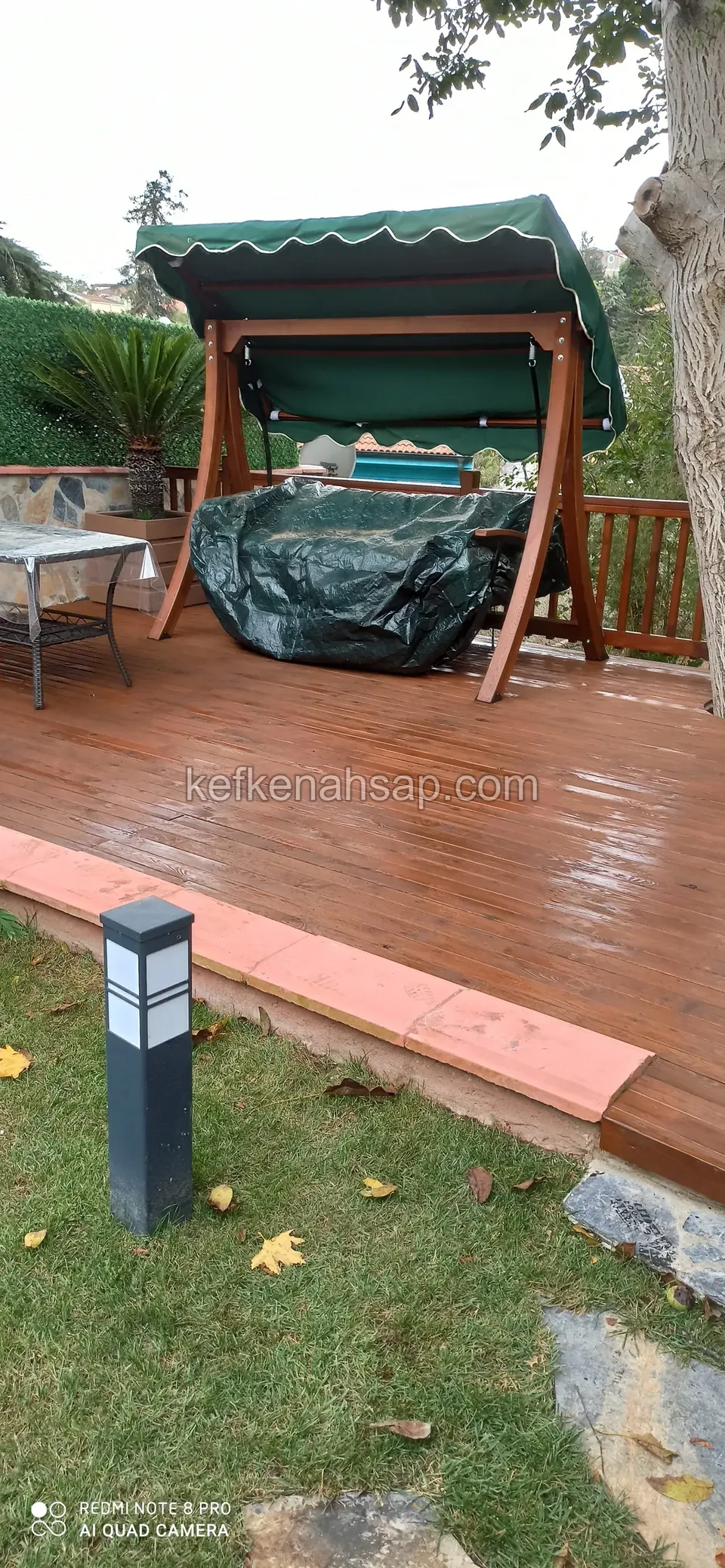 Kırmızımsı boyalı ahşap deck üzerinde yeşil tenteli bahçe salıncağı, kenarda bahçe lambası