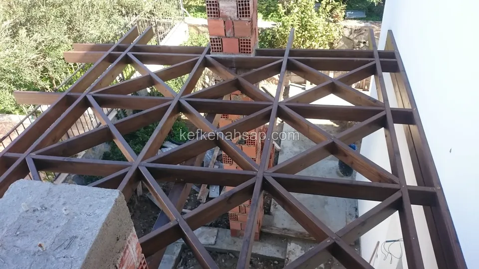 Yukarıdan görünen geometrik pergola kafesi, tuğla kolonlar, beyaz duvar ve çevrede çalılık ile zeytin ağaçları