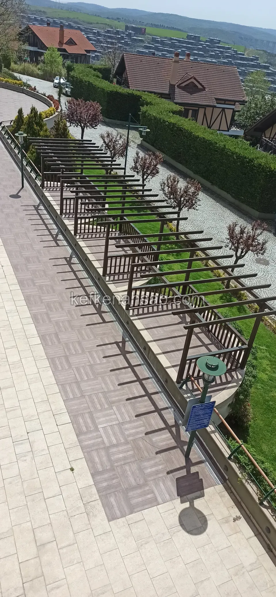 Lange Reihe aus Holzpergola und Geländer entlang eines Weges von oben gesehen, mit Rasen und Häusern in der Ferne