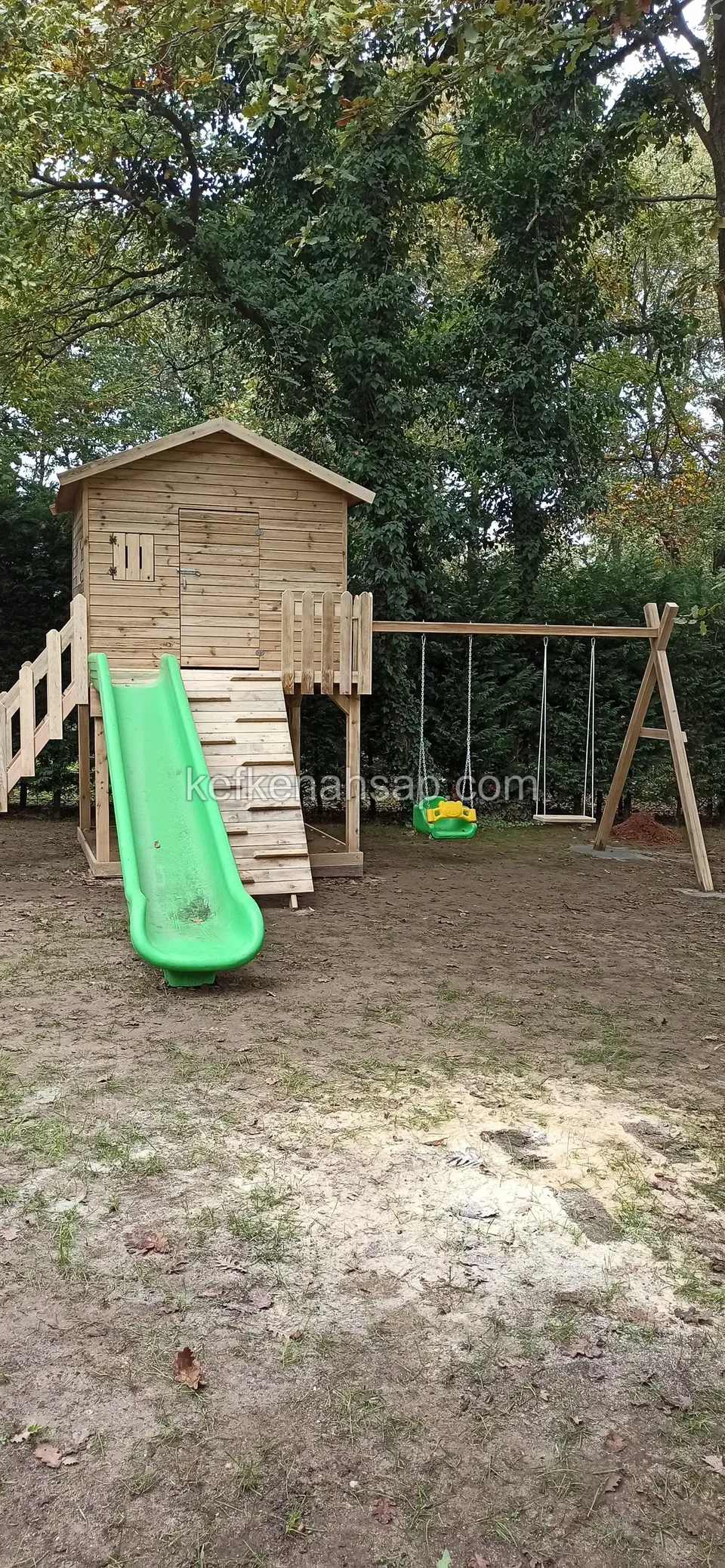 Toprak zeminde duran yüksek ahşap oyun evi, yeşil kaydırağı ve yan tarafta salıncağı
