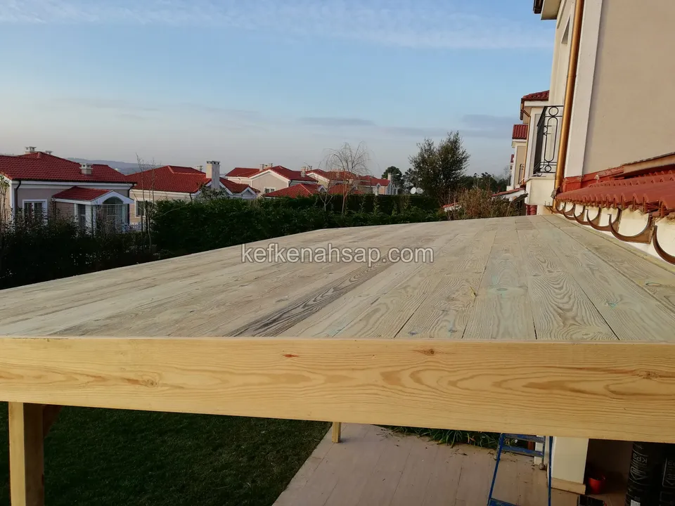 Edge of a raised timber deck with light boards, with houses and a hedge behind