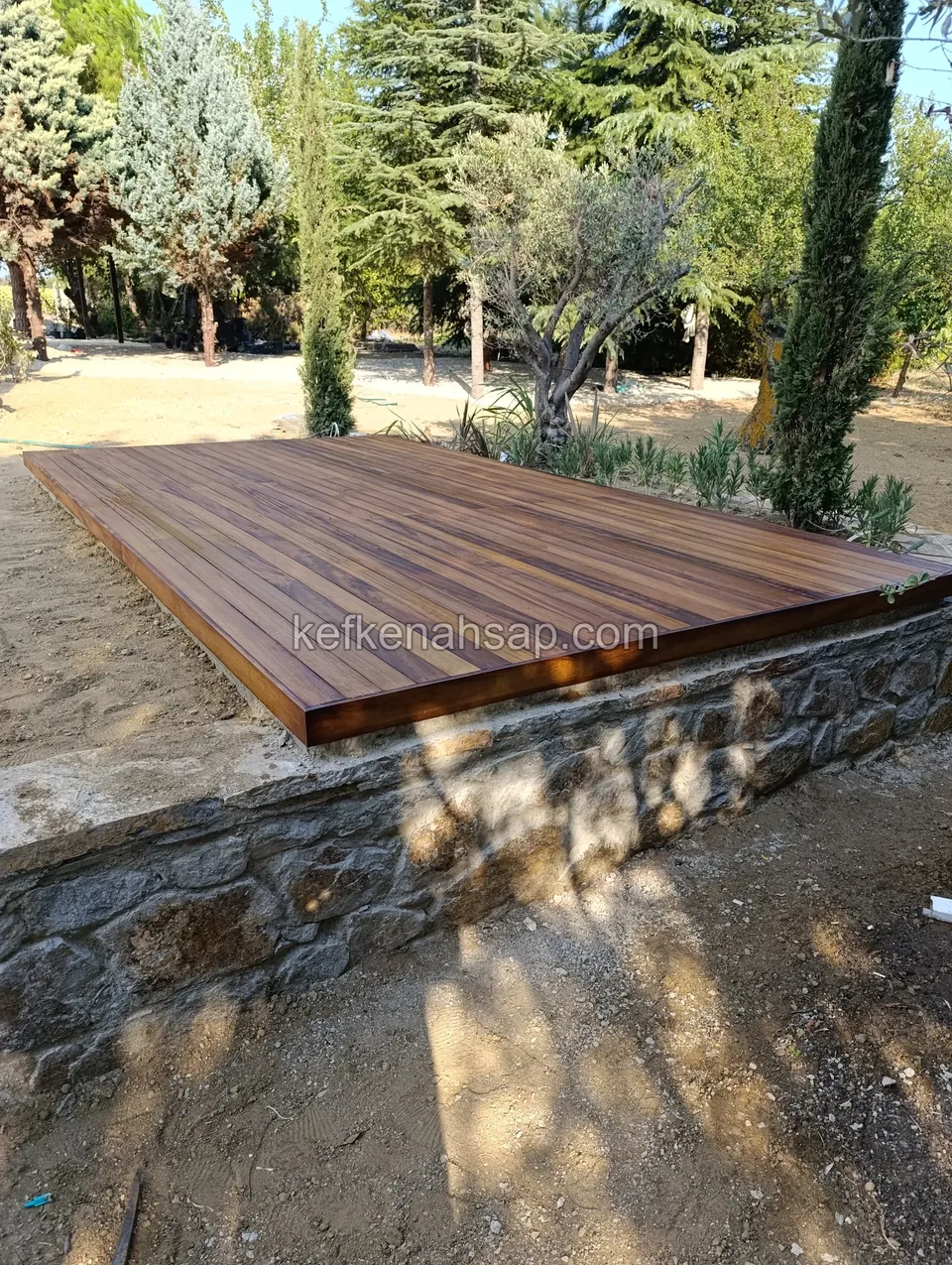 Dark-toned timber deck lying on top of a stone wall rising from sandy ground, with trees and a slim cypress behind