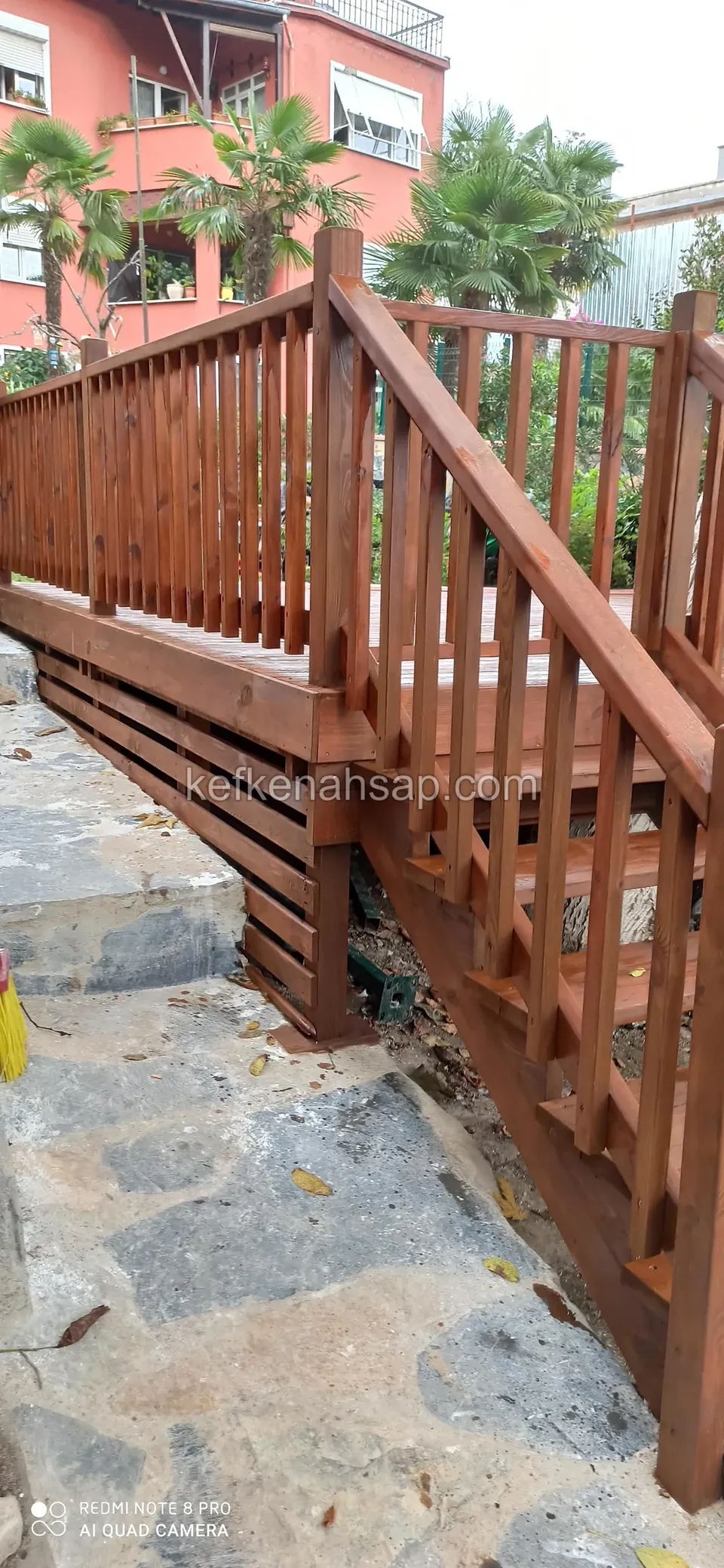 درابزين خشبي ودرابزين درج لديكينغ مرتفع مطلي بالبني المحمرّ بجانب أرضية حجرية، وخلفه نخيل ومبنى