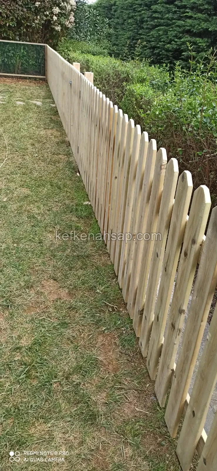 Çim alanın kenarında, arkasında çalılık bulunan, yuvarlak başlı dikey çıtalardan yapılmış açık renkli ahşap bahçe çiti