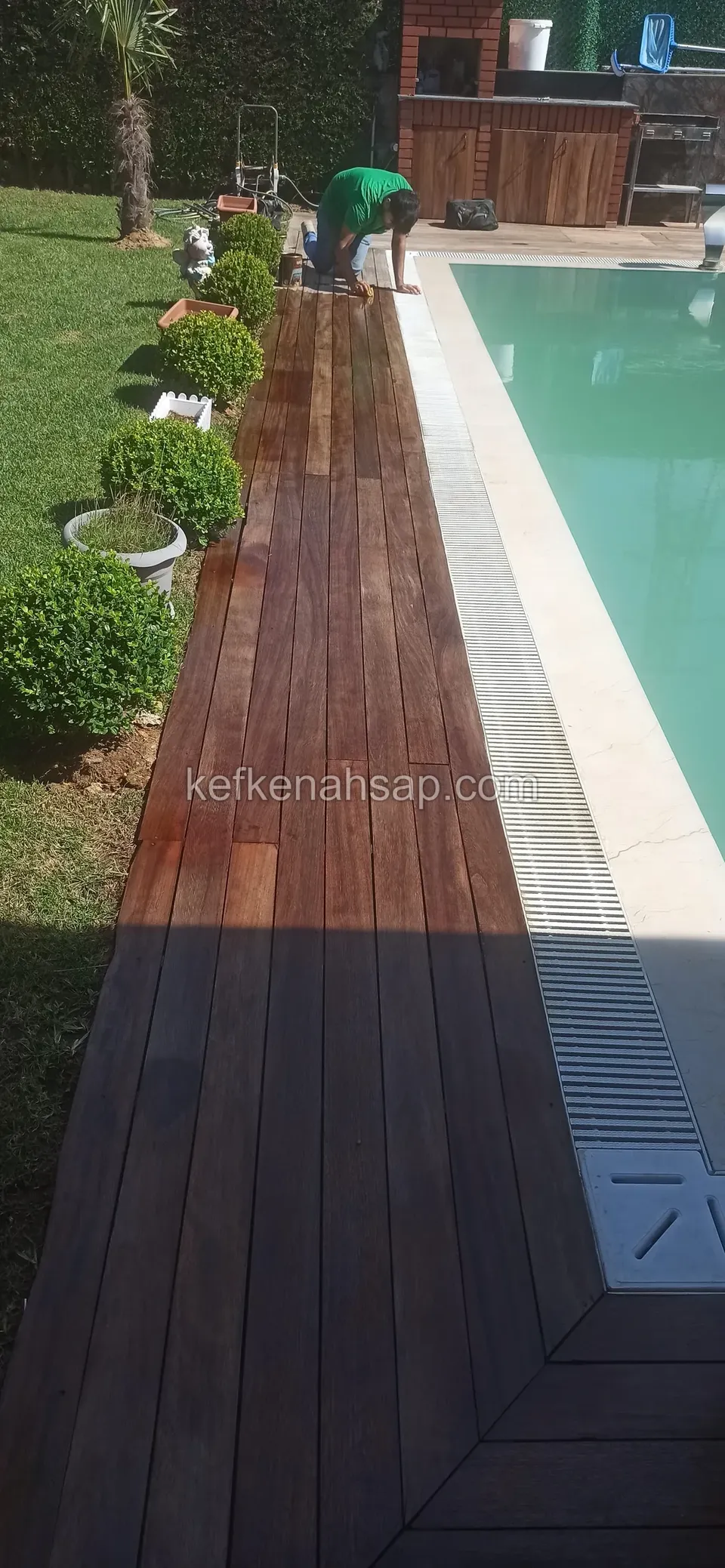 Dark brown deck strip along the grated edge of a pool, with round-clipped shrubs on its left