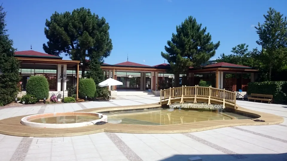 Kırmızı çatılı yapıların önünde, ahşap deck ile çevrili havuz ve uzak uçtaki kemerli köprü