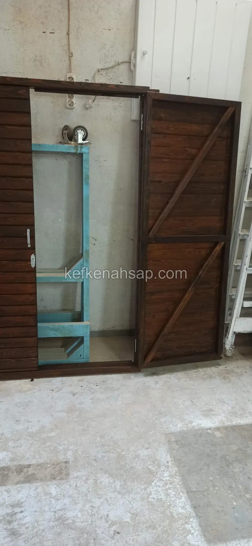Koyu ahşap dolabın Z çaprazlı kapak kanadı açık, içinde açık mavi boyalı bir çerçeve ve duvarda ampul