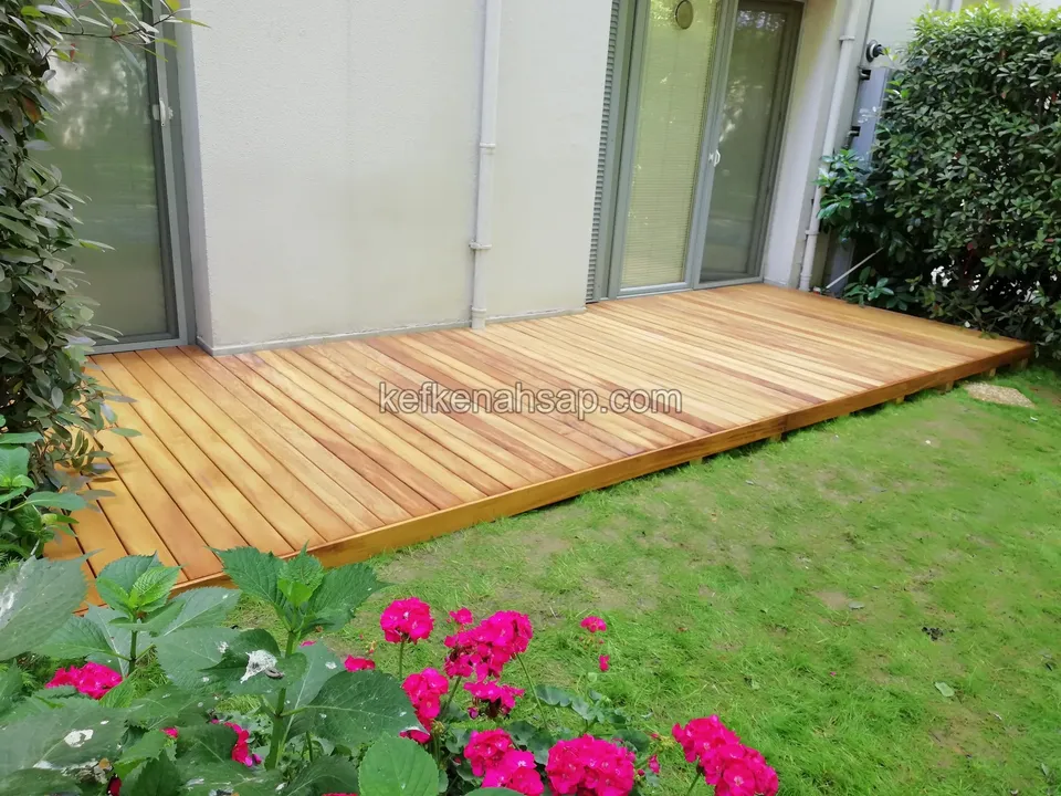 Golden-brown timber deck laid in front of a ground-floor flat's lawn, with pink flowers in front