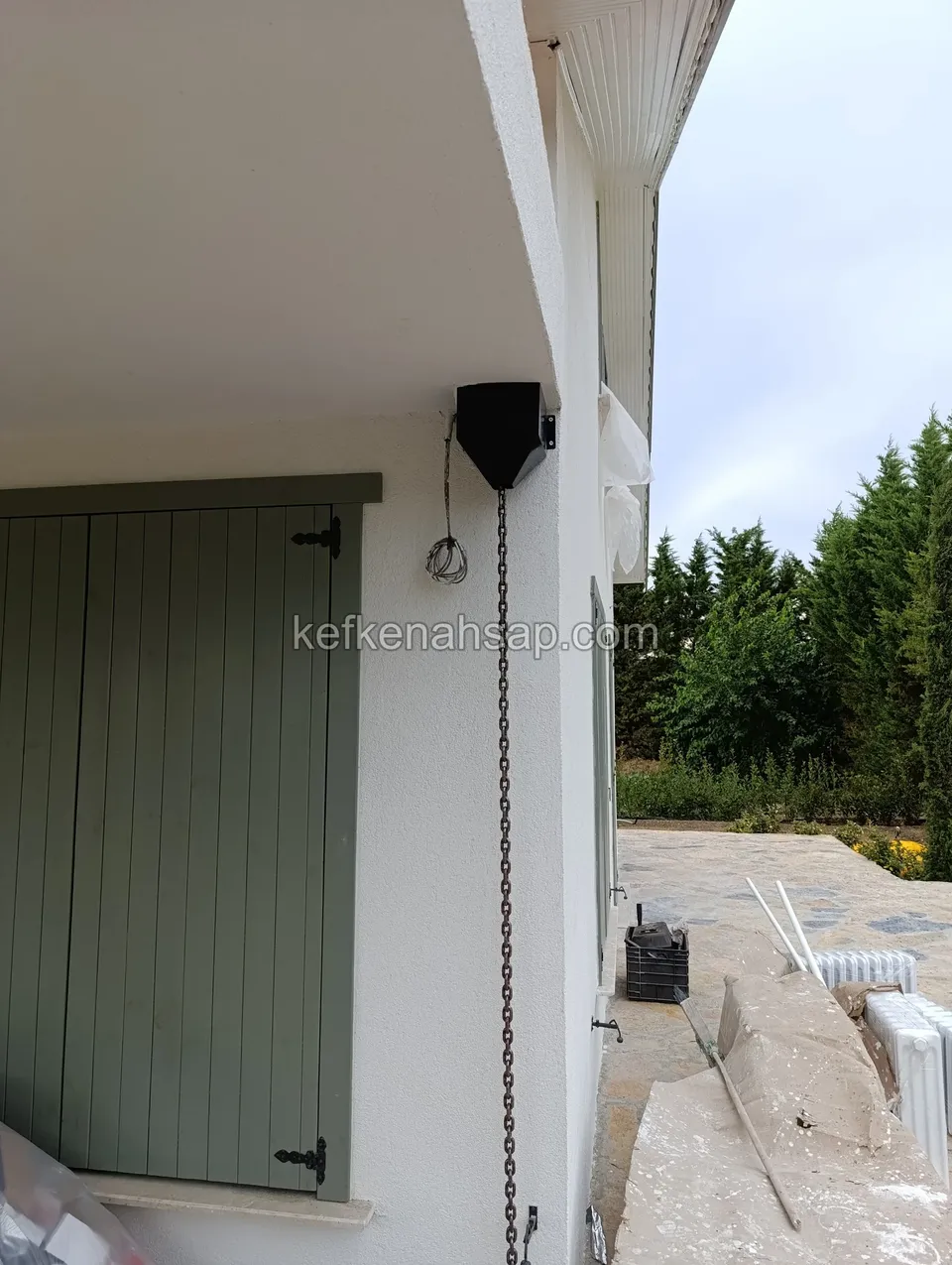 Beyaz cephede kapalı duran yeşil dikey çıtalı panjur kanadı, siyah menteşeler ve yanında zincir sarkan bir mekanizma