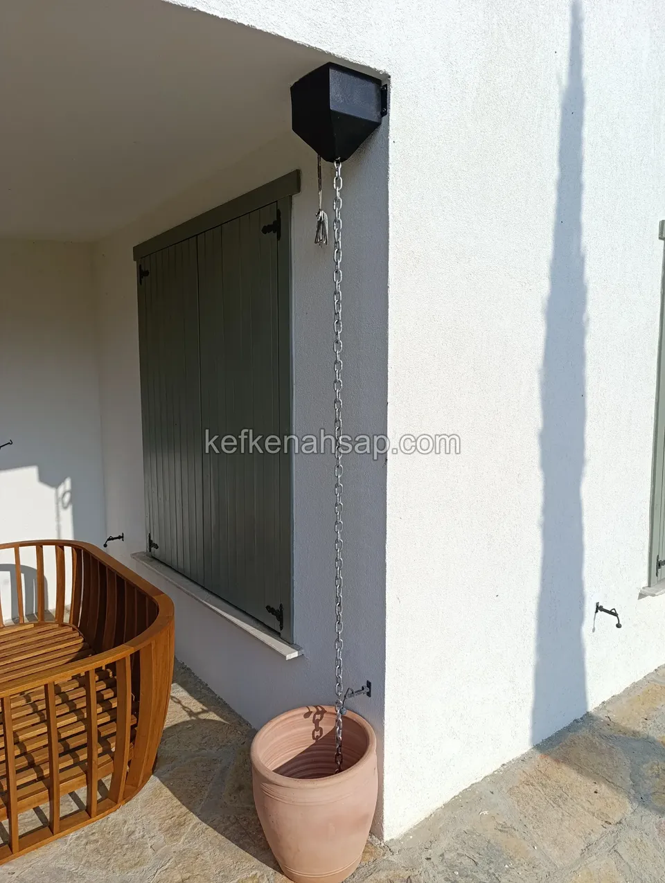 Beyaz cephenin köşesinde, kapalı adaçayı yeşili panjurun yanında çatıdan sarkan zincir, terracotta saksı ve ahşap bir koltuk